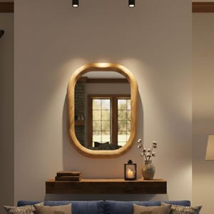 Peut inclure: Un miroir mural ovale avec un cadre en bois clair monté au-dessus d'une étagère flottante en bois brun foncé. L'étagère supporte une pile de livres, une lanterne noire avec une bougie blanche et un vase contenant des tiges de coton.