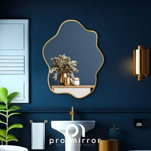 May include: A gold-framed, irregular-shaped mirror hangs on a dark blue wall in a bathroom setting. The mirror reflects a bouquet of dried flowers in a gold vase, along with other decorative items on a shelf.