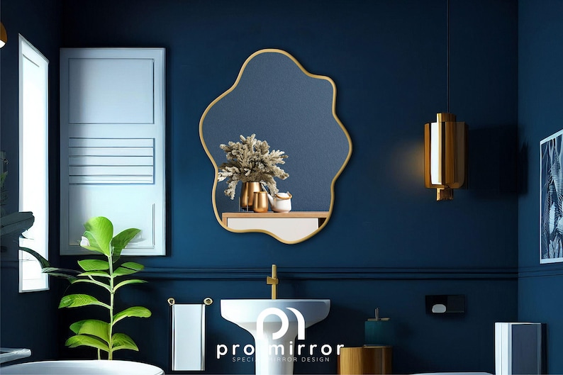 May include: A gold-framed, irregular-shaped mirror hangs on a dark blue wall in a bathroom setting. The mirror reflects a bouquet of dried flowers in a gold vase, along with other decorative items on a shelf.