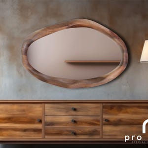 May include: A wooden oval mirror with an irregular, organic shape hangs on a textured wall above a wooden dresser with three drawers. The mirror has a light brown wood frame and a reflective surface. The dresser has a light brown finish and black drawer pulls.