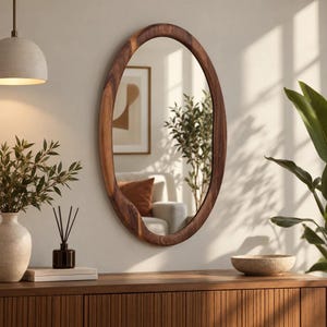 May include: An oval wall mirror with a thick, natural dark brown wood frame featuring a visible grain pattern. The mirror reflects a room interior with a sofa, a framed wall art piece, and a tall green plant, showcasing a smooth, polished surface.