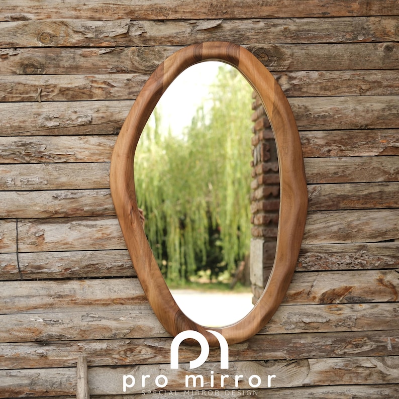 May include: A wooden oval mirror with an irregular, organic shape. The mirror is hanging on a wooden wall and reflects a green tree and a brick wall.