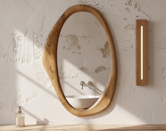 May include: An oval mirror with a thick, natural wood frame featuring a prominent grain pattern. To the right, a rectangular wooden wall light fixture with a vertical LED strip. The mirror reflects a white basin sink and silver faucet hardware.