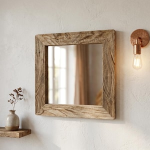 May include: Square mirror with a rustic wooden frame. The mirror is mounted on a white wall, with a copper-coloured wall sconce to the right. A small shelf with a vase and dried flowers is to the left.