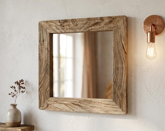 May include: Square wooden framed mirror with a natural, weathered finish. The mirror is mounted on a white wall, with a copper-colored wall sconce to the right. A small shelf with a vase and dried flowers is to the left.