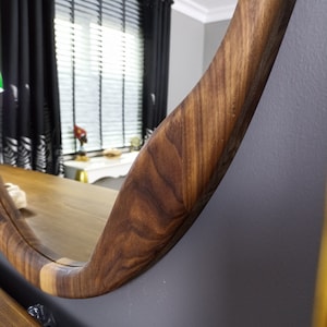 May include: A circular wall mirror featuring a thick, polished wooden frame with a prominent natural grain pattern. The wood displays varying shades of brown, showcasing a smooth, curved edge that reflects the surrounding room in its glass surface.