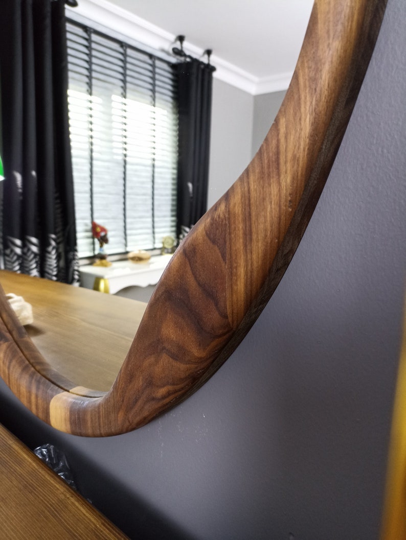 May include: A circular wall mirror featuring a thick, polished wooden frame with a prominent natural grain pattern. The wood displays varying shades of brown, showcasing a smooth, curved edge that reflects the surrounding room in its glass surface.