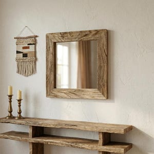 May include: Square wooden framed mirror with a natural, weathered finish. A woven wall hanging with beige, black, and brown patterns hangs to the left. Two brass candle holders with lit candles sit on a wooden console.