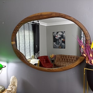 May include: An oval wall mirror with a natural wood frame featuring an irregular, organic edge. The reflective surface shows a room interior with a red armchair, a brown tufted sofa, a wall art piece, and a window with dark blinds.