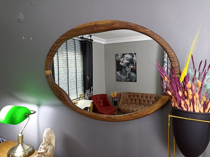 May include: An oval wall mirror with a natural wood frame featuring an irregular, organic edge. The reflective surface shows a room interior with a red armchair, a brown tufted sofa, a wall art piece, and a window with dark blinds.