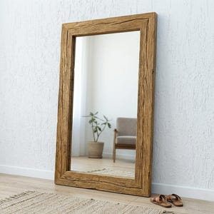 Può includere: Un grande specchio rettangolare a figura intera con una spessa cornice in legno strutturato. Lo specchio è appoggiato a una parete bianca, che riflette una stanza con una sedia, una pianta e un tappeto. Sandali marroni sul pavimento.