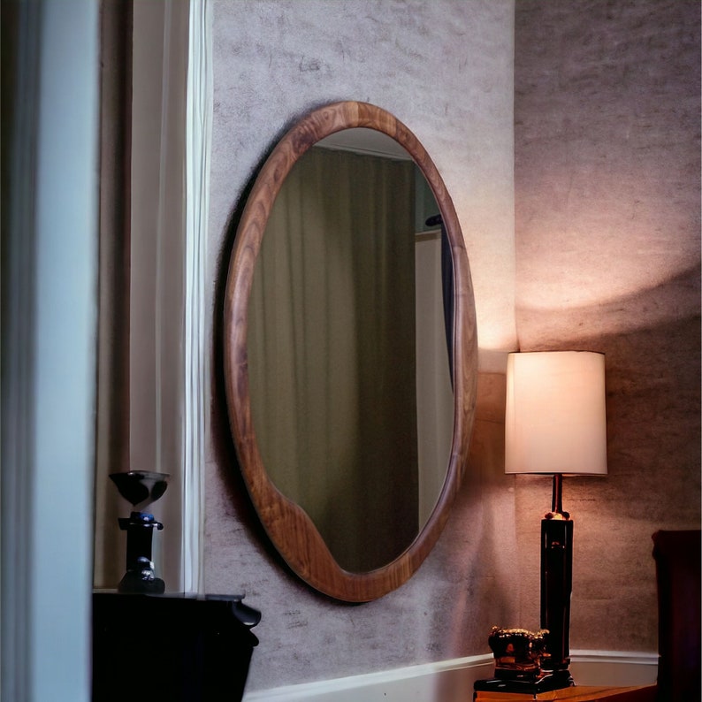 May include: A large, oval-shaped wall mirror featuring a thick, natural-toned wooden frame with an organic, slightly irregular edge. The mirror surface is clear and reflective, capturing the surrounding room environment within its polished glass.