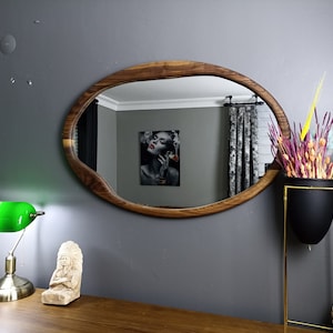 May include: An oval wall mirror with a natural wood frame featuring an irregular, organic edge. The mirror reflects a room interior with a wall art piece showing a person with red lips. The frame displays a distinct wood grain pattern throughout.