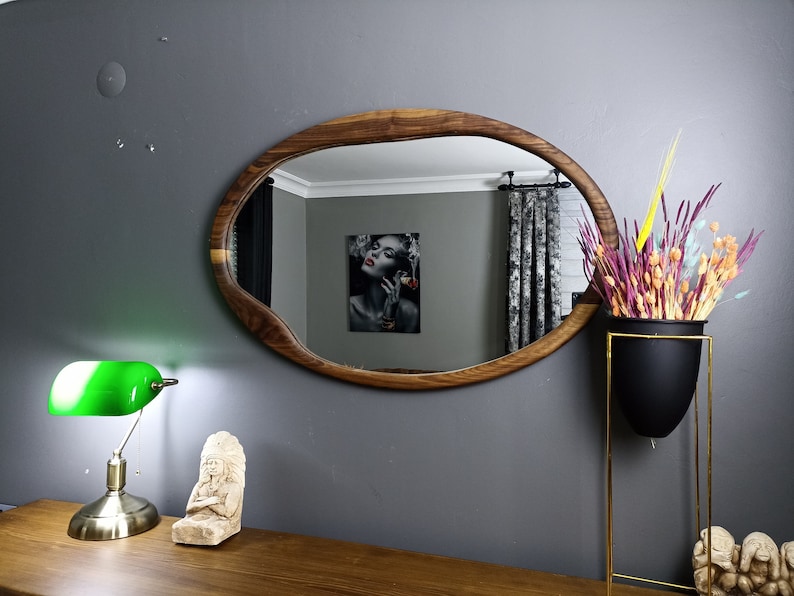 May include: An oval wall mirror with a natural wood frame featuring an irregular, organic edge. The mirror reflects a room interior with a wall art piece showing a person with red lips. The frame displays a distinct wood grain pattern throughout.