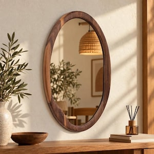 May include: An oval wall mirror featuring a thick, dark brown wooden frame with a visible natural grain pattern. The mirror reflects a room interior, including a woven pendant light, a potted plant, and a framed picture on the wall.