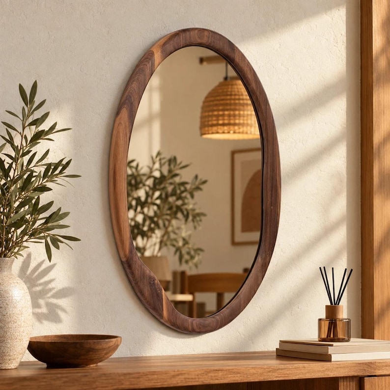 May include: An oval wall mirror featuring a thick, dark brown wooden frame with a visible natural grain pattern. The mirror reflects a room interior, including a woven pendant light, a potted plant, and a framed picture on the wall.