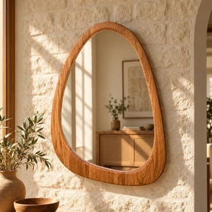 Peut inclure: Un miroir mural doté d'un cadre en bois irrégulier et organique en forme de galet, avec un grain de bois brun clair visible. La surface réfléchissante montre l'intérieur d'une pièce avec une armoire en bois et une plante.