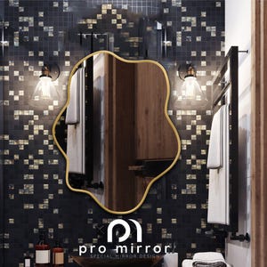 May include: A gold-framed, wavy-shaped mirror hangs on a black and gold mosaic tile wall in a bathroom. The mirror reflects a wooden cabinet and a white towel hanging on a black towel rack. The text "pro mirror SPECIAL MIRROR DESIGN" is visible below the mirror.