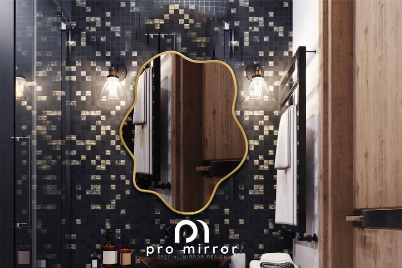 May include: A gold-framed, wavy-shaped mirror hangs on a black and gold mosaic tile wall in a bathroom. The mirror reflects a wooden cabinet and a white towel hanging on a black towel rack. The text "pro mirror SPECIAL MIRROR DESIGN" is visible below the mirror.