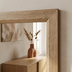 Peut inclure: Un miroir rectangulaire dot&eacute; d'un cadre &eacute;pais en bois de ch&ecirc;ne clair au grain apparent. Le miroir refl&egrave;te un vase en c&eacute;ramique brun clair &agrave; la base arrondie en forme d'arche, contenant des tiges d'herbe de la pampa s&eacute;ch&eacute;es.