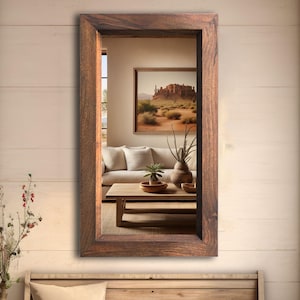 May include: A rectangular, rustic wooden framed mirror. The mirror reflects a room with a sofa, coffee table, and a painting of a desert landscape. The frame has a dark brown finish.