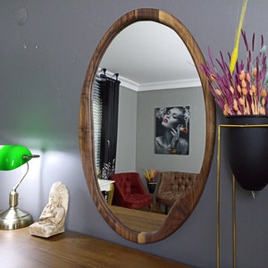 May include: An oval wall mirror with a natural dark brown wood frame. The mirror reflects a room interior containing a red armchair, a brown armchair, and a framed wall print. The mirror is mounted on a gray wall above a wooden surface.