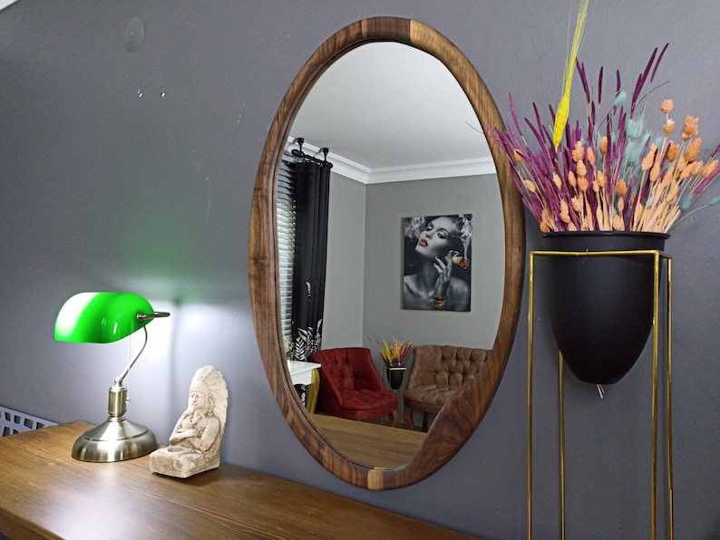 May include: An oval wall mirror with a natural dark brown wood frame. The mirror reflects a room interior containing a red armchair, a brown armchair, and a framed wall print. The mirror is mounted on a gray wall above a wooden surface.