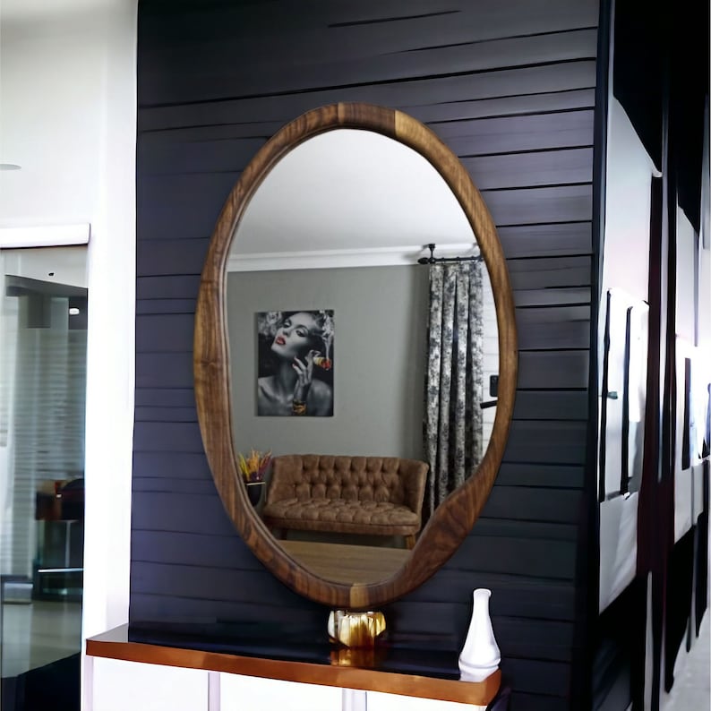 May include: An oval-shaped mirror with a natural wood grain frame. The mirror reflects a room containing a brown tufted sofa, a wall art piece featuring a person, and patterned curtains. The mirror is positioned above a console table.