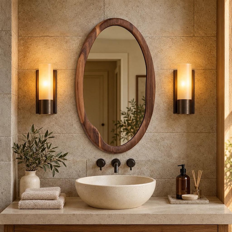 May include: An oval mirror with a dark brown natural wood frame mounted above a beige stone vessel sink. The sink features a dark metal faucet and two matching wall-mounted handles. Two cylindrical wall sconces with warm light flank the mirror.