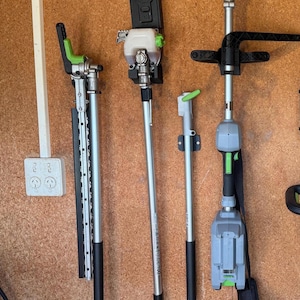 May include: Four different types of gardening tools hanging on a wall. The tools are all black and green and have long silver poles. The tools are labelled "EGO" and "DEWALT".
