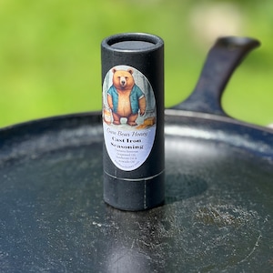 May include: A black tube of cast iron seasoning with a cartoon bear wearing a blue shirt and holding a jar of honey. The label reads "Gare Bear Honey Cast Iron Seasoning" and lists the ingredients: beeswax, grapeseed oil, sunflower oil, and avocado oil. The tube is sitting on a black cast iron skillet.