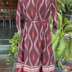 May include: A long, maroon dress with a geometric pattern in shades of red, pink, and white. The dress features long sleeves, a mandarin collar, and a matching belt. The hemline has a decorative border.