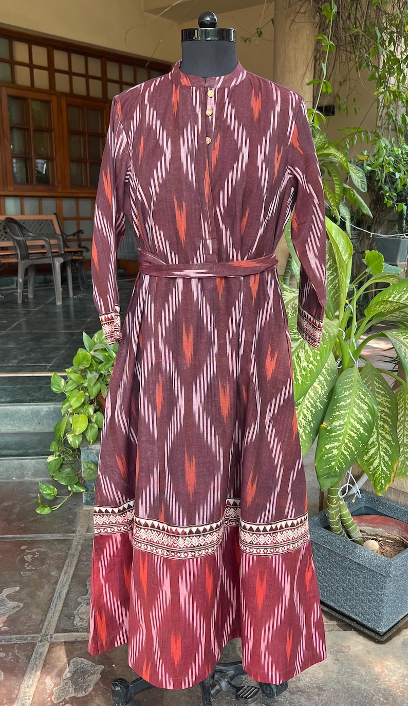 May include: A long, maroon dress with a geometric pattern in shades of red, pink, and white. The dress features long sleeves, a mandarin collar, and a matching belt. The hemline has a decorative border.
