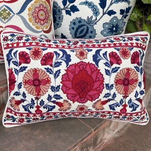 May include: A rectangular decorative pillow with a white background and a floral pattern in shades of red, pink, and blue. The pillow has a white border and is part of a set of pillows with similar designs. The pillow is likely made of a soft fabric.