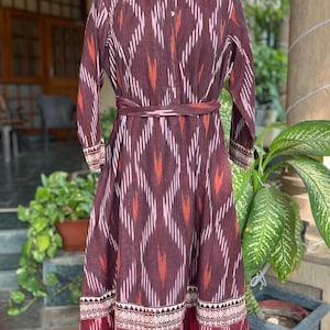 May include: A long, maroon dress with a geometric pattern in white, orange, and pink. The dress features long sleeves, a mandarin collar, and a matching belt. The hemline and cuffs have a decorative border.