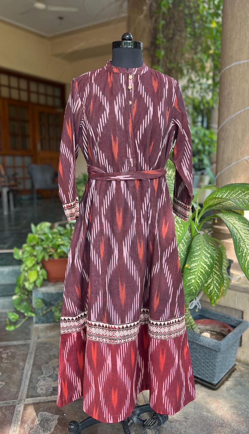 May include: A long, maroon dress with a geometric pattern in white, orange, and pink. The dress features long sleeves, a mandarin collar, and a matching belt. The hemline and cuffs have a decorative border.