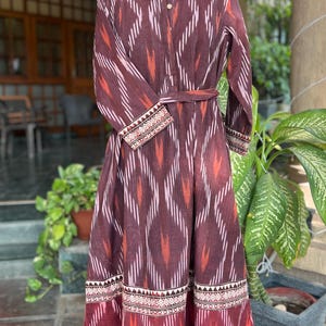 May include: A long, maroon dress with a geometric pattern in white and orange. The dress features a mandarin collar, long sleeves, and a matching belt. The hem and cuffs are detailed with a patterned border. The dress is displayed on a mannequin.