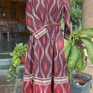 May include: A long, maroon dress with a geometric ikat pattern in white, orange, and pink. The dress features long sleeves, a mandarin collar, and a matching belt. The hem and cuffs have a decorative border.