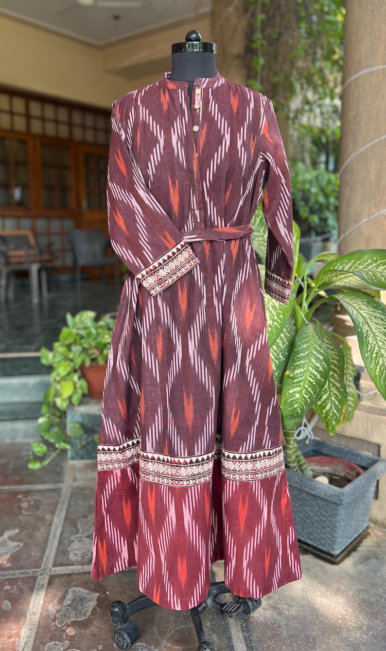 May include: A long, maroon dress with a geometric ikat pattern in white, orange, and pink. The dress features long sleeves, a mandarin collar, and a matching belt. The hem and cuffs have a decorative border.