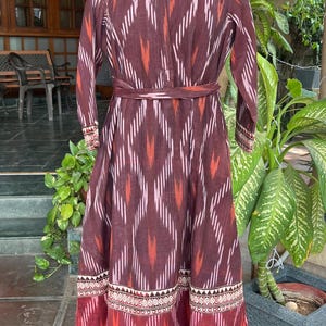May include: A long, maroon dress with a geometric pattern in white, orange, and pink. The dress features long sleeves, a mandarin collar, and a matching belt. The hemline and cuffs have a decorative border.