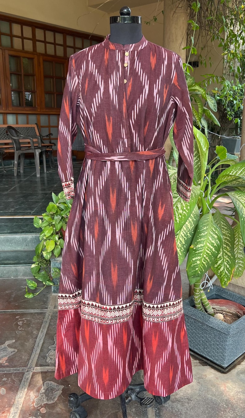 May include: A long, maroon dress with a geometric pattern in white, orange, and pink. The dress features long sleeves, a mandarin collar, and a matching belt. The hemline and cuffs have a decorative border.