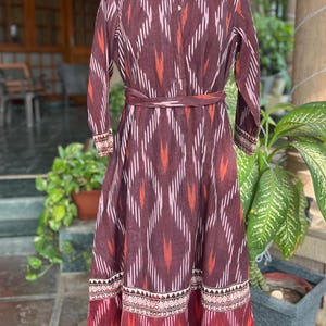 May include: A long, maroon dress with a geometric ikat pattern in white, orange, and pink. The dress features long sleeves, a mandarin collar, and a matching belt. The hemline and cuffs have a decorative border.