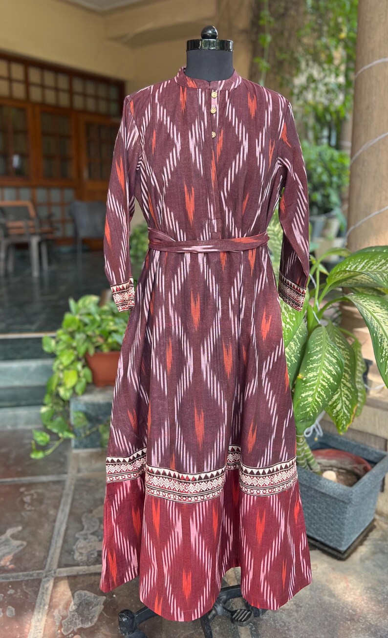 May include: A long, maroon dress with a geometric ikat pattern in white, orange, and pink. The dress features long sleeves, a mandarin collar, and a matching belt. The hemline and cuffs have a decorative border.