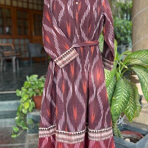 May include: A long, maroon dress with a geometric pattern in white and orange. The dress features long sleeves, a mandarin collar, and a matching belt. The hem and cuffs are detailed with a patterned border. The dress is displayed on a mannequin.