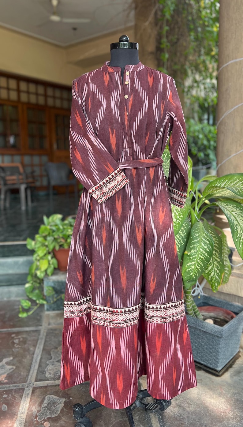 May include: A long, maroon dress with a geometric pattern in white and orange. The dress features long sleeves, a mandarin collar, and a matching belt. The hem and cuffs are detailed with a patterned border. The dress is displayed on a mannequin.
