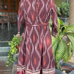 May include: A long, maroon dress with a geometric pattern of white and orange stripes. The dress has long sleeves, a mandarin collar, and a matching belt. The hemline features a decorative border.