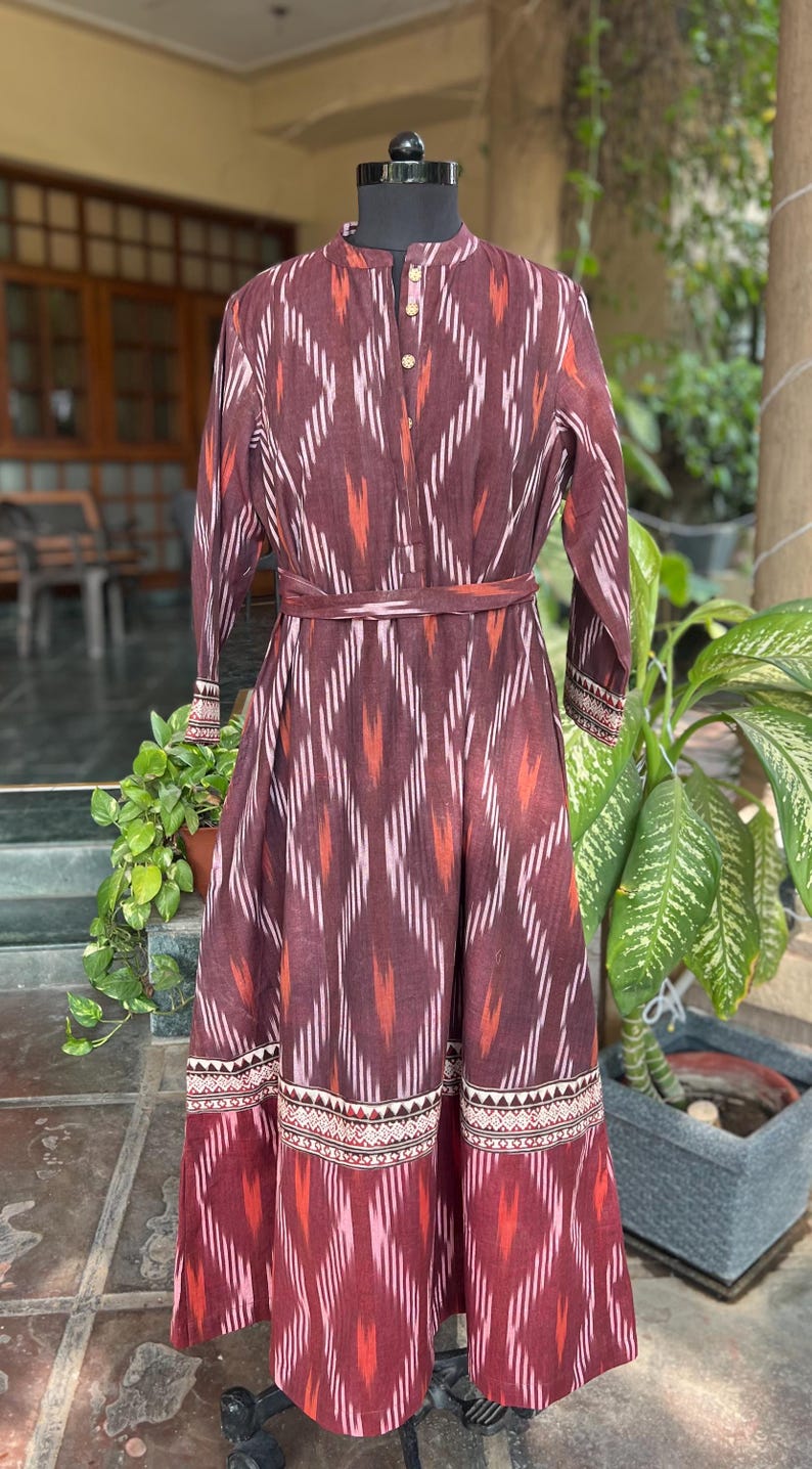 May include: A long, maroon dress with a geometric pattern of white and orange stripes. The dress has long sleeves, a mandarin collar, and a matching belt. The hemline features a decorative border.