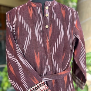 May include: A long-sleeved, burgundy dress with a mandarin collar and a matching belt. The dress features a geometric ikat pattern in white and orange. The cuffs are adorned with a detailed, embroidered design in red, white, and black.