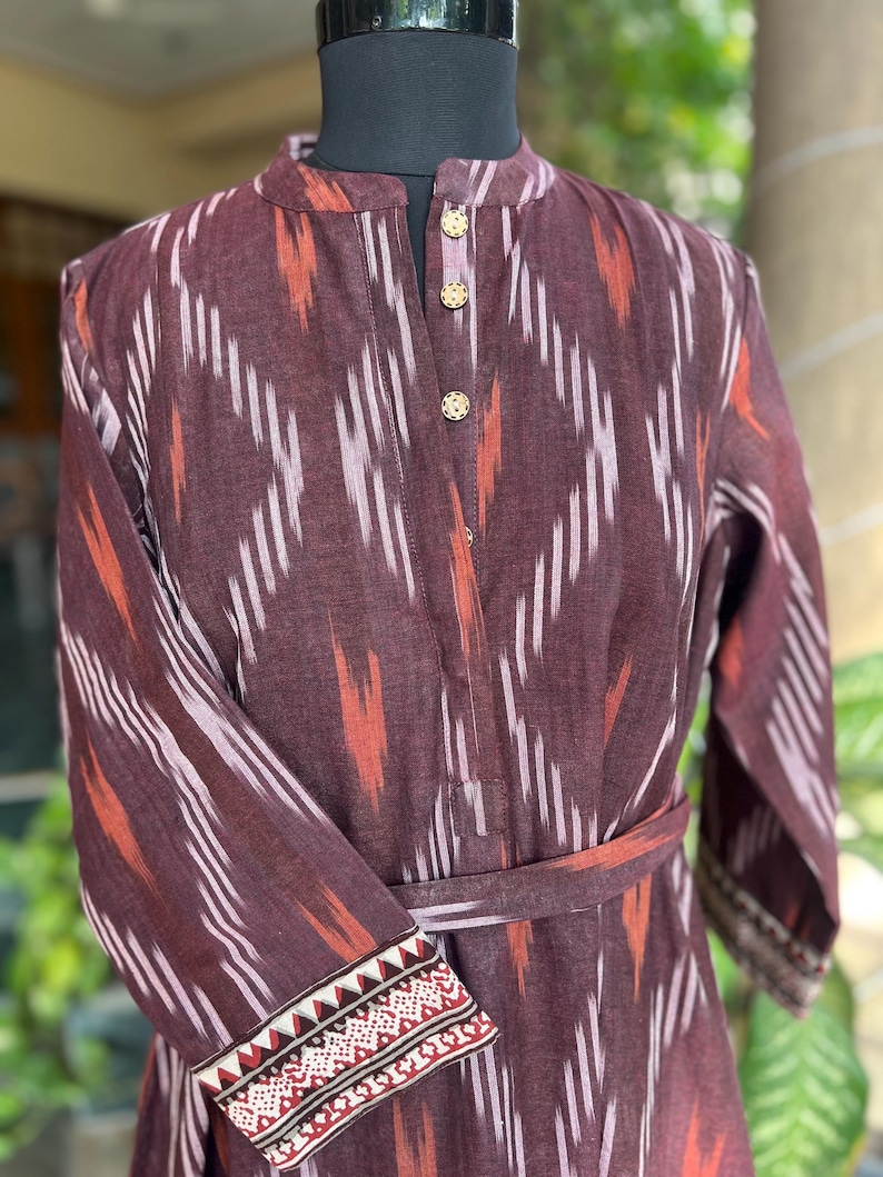 May include: A long-sleeved, burgundy dress with a mandarin collar and a matching belt. The dress features a geometric ikat pattern in white and orange. The cuffs are adorned with a detailed, embroidered design in red, white, and black.