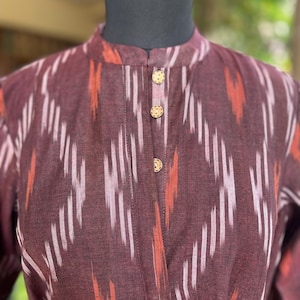 May include: A maroon dress with a mandarin collar and three gold buttons. The fabric features a white and orange ikat pattern. The dress is displayed on a black mannequin head.